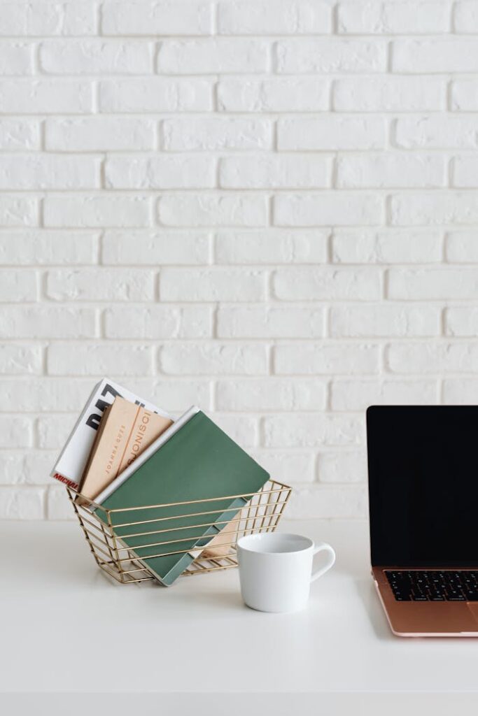 A clean and organized desk setup with a laptop, cup, and notebooks against a white brick wall.